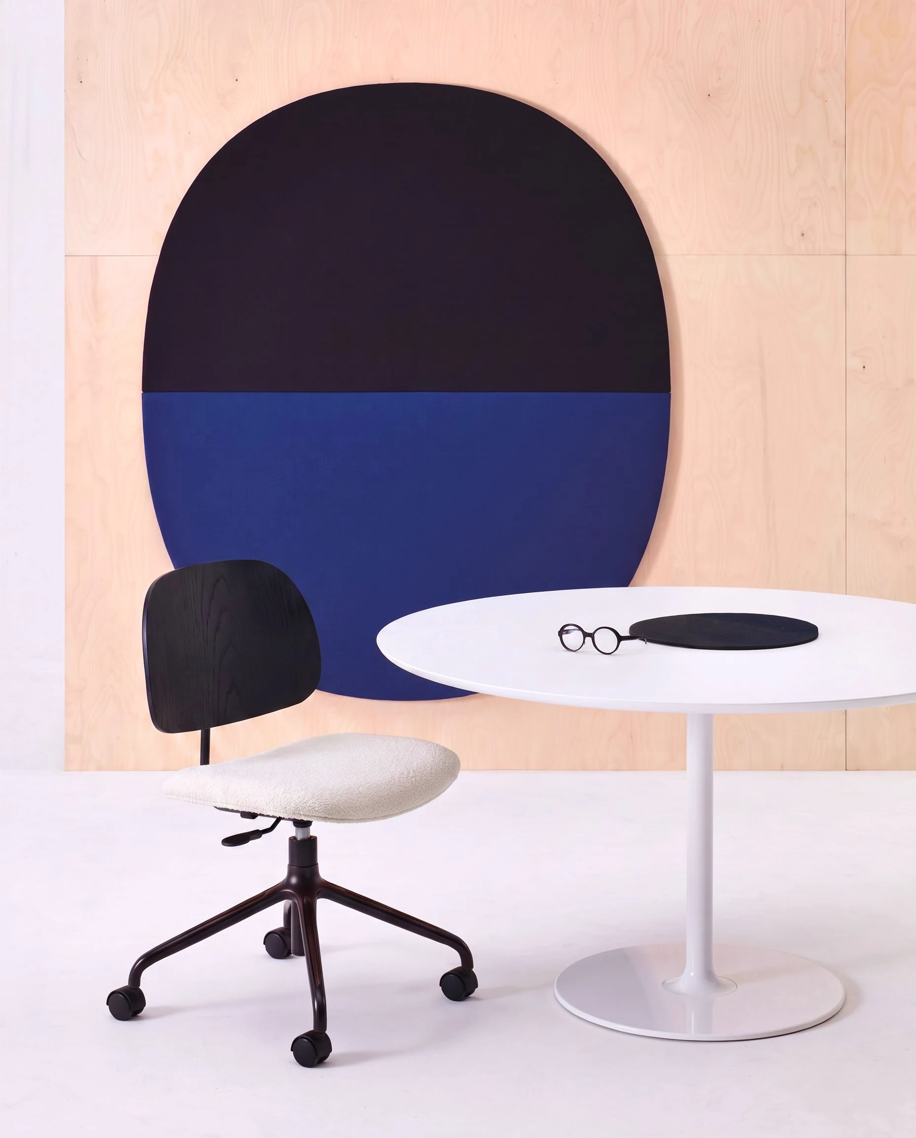 Cadeira Nômia Office preta com assento estofado junto de mesa redonda branca, diante de painel em arco azul-marinho